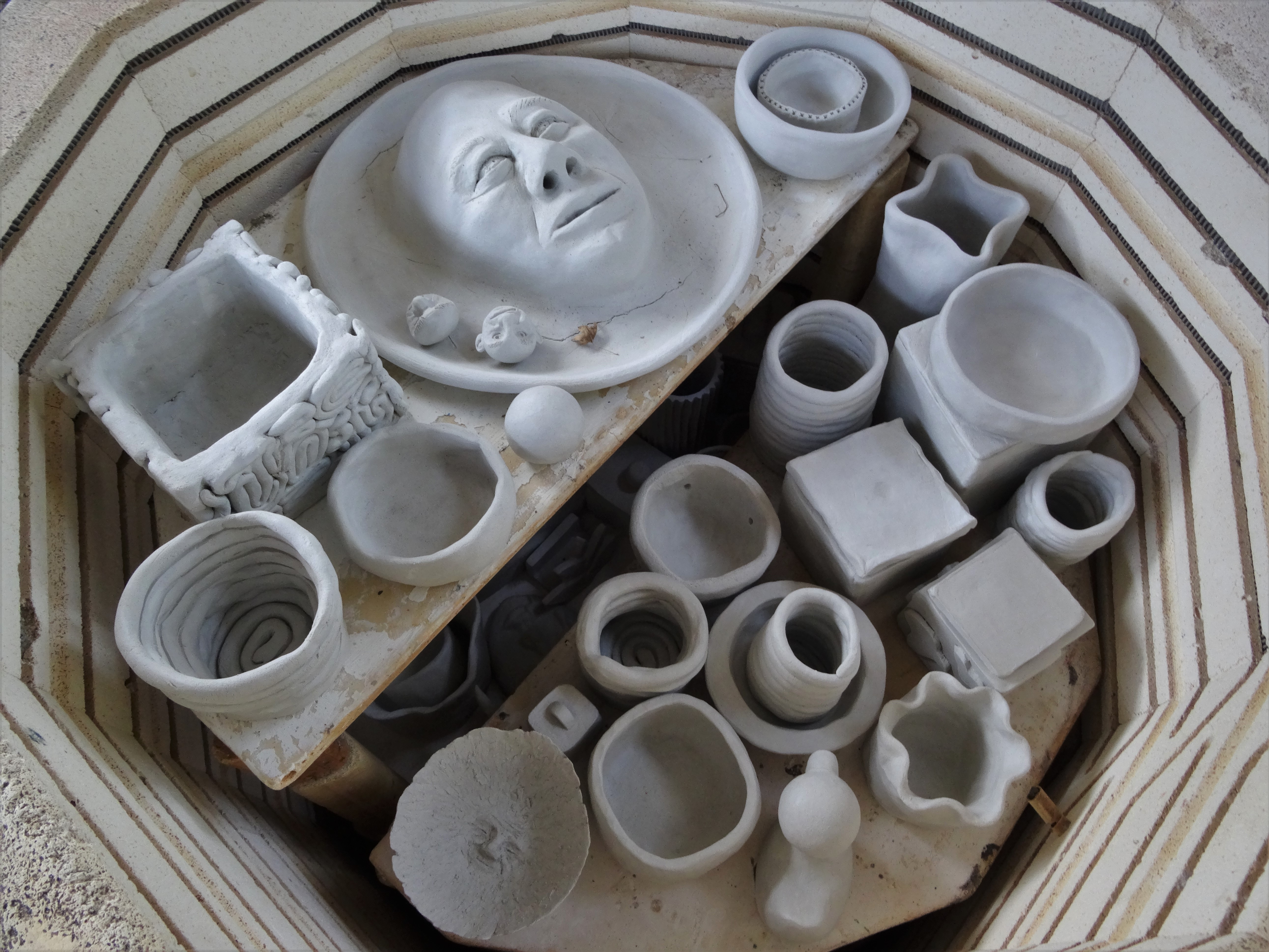 A variety of scultures such as a plate with a face and many vases and bowls fill a kiln, ready to be fired. 