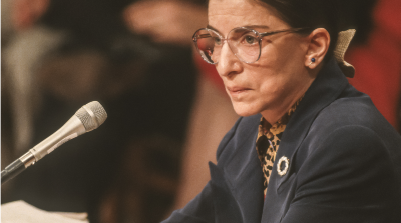 Justice Ruth Bader Ginsburg sitting with arms crossed in front of a microphone