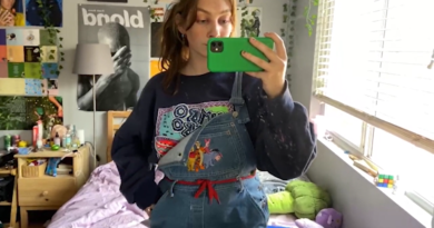 Susannah Costello taking a mirror selfie in her room with her green-cased iPhone wearing a navy blue sweater and overalls