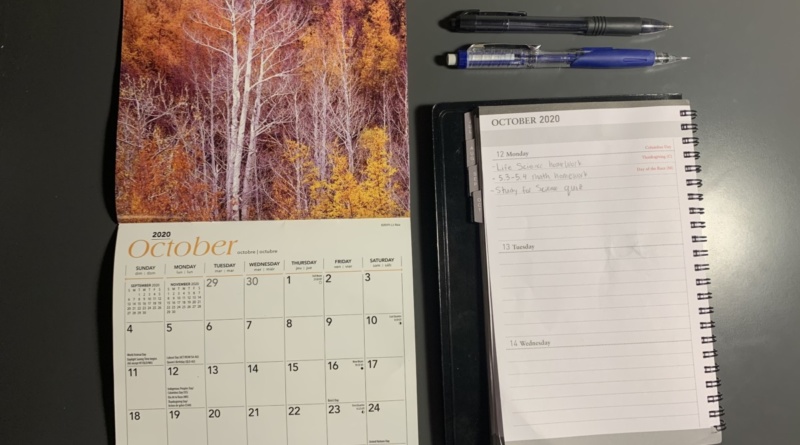 A calendar and a planner both opened to the month of October are splayed out on a desk along with a black gel pen and blue mechanical pencil