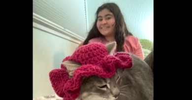Olivet Yettou poses smiling with one of her cats, who is wearing a maroon, creased cap.