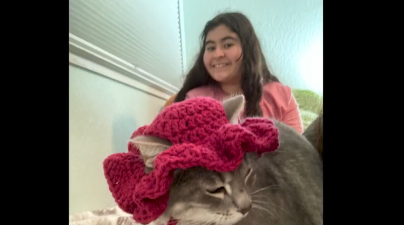 Olivet Yettou poses smiling with one of her cats, who is wearing a maroon, creased cap.