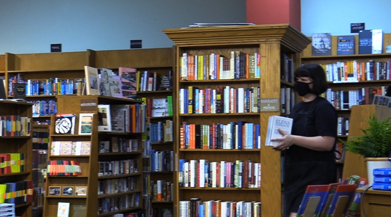 Books fill the shelves inside the Avid Reader bookstore. A customer wearing a mask stands on the right holding a book
