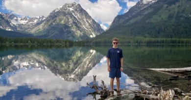 Luca Maes standing on a rock with sunglasses on. The mountains and clouds reflect off the water of the lake behind him.