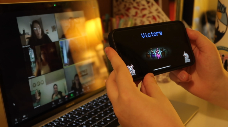A student holding up a phone with the victory screen from the game Among Us in front of a Zoom meeting on a laptop