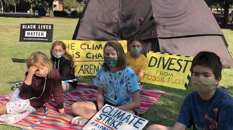 Five kids wearing face masks sit in the grass on a blanket. They are in front of a tent which insinuates that they are camping out and they are holding climate activism signs that say things like "divest from fossil fules" and "climate strike, Fridays 12-1 central park"